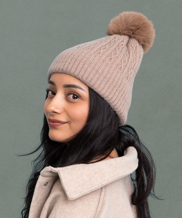 Woman wearing a beige knit beanie with a pom-pom against a gray background | Three Mad Hatters Mercantile